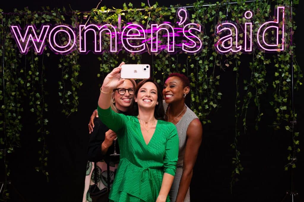 Three women taking a picture underneath a light-up Women's Aid sign