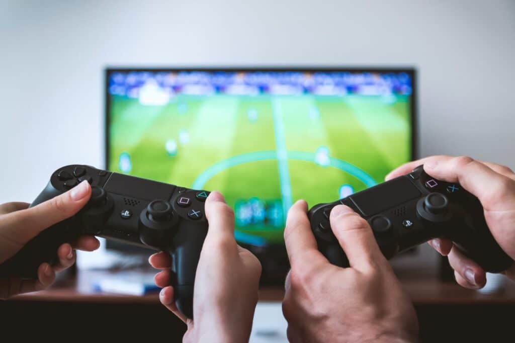 Hands holding PlayStation controllers with a tv in the background