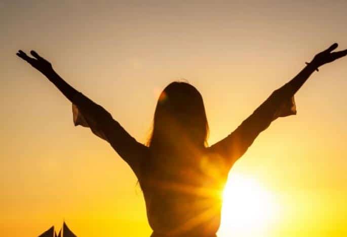 Silhouette of woman standing holding her arms up with the sun shining on her